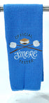 A blue microfiber waffle weave hand towel with 'OFFICIAL SMORE TESTER' embroidered on it, along with an image of a smore and marshmallows on sticks.